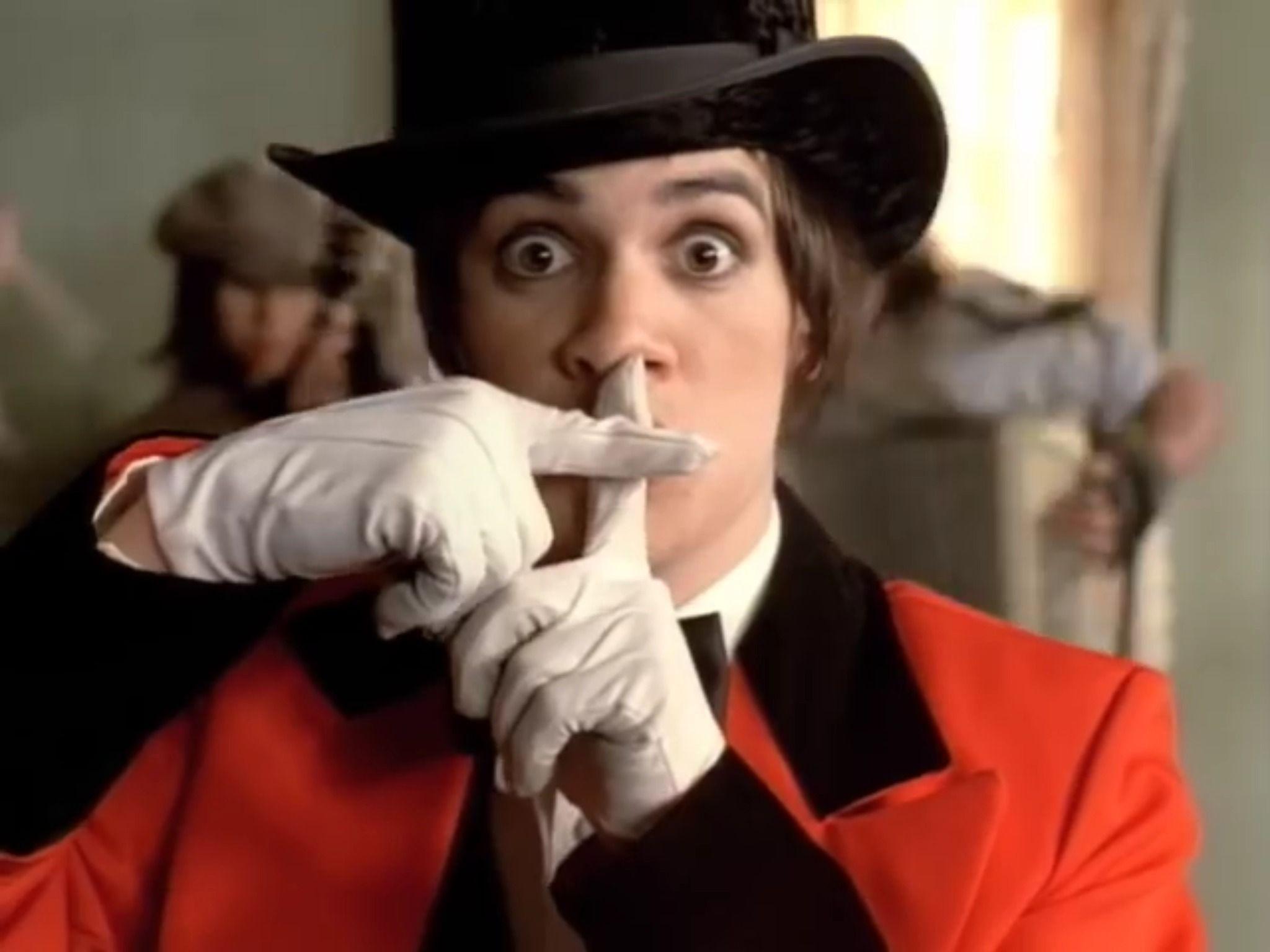 A screenshot from the music video for Panic! at the Disco's 'I Write Sins, Not Tragedies' featuring a wide-eyed man in a black top hot and red and black suit making a cross with white-gloved fingers in front of his mouth.